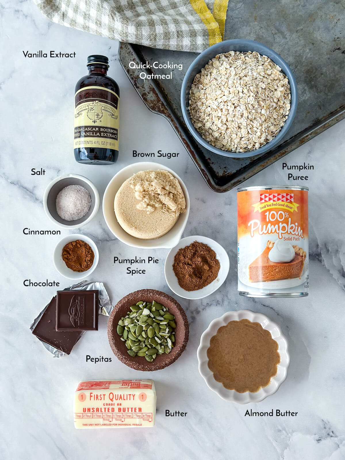 Ingredients for no bake pumpkin cookies on a marble surface including: vanilla, quick-cooking oatmeal, brown sugar, pumpkin puree, salt, cinnamon, chocolate, pumpkin pie spice, pepitas, butter and almond butter.