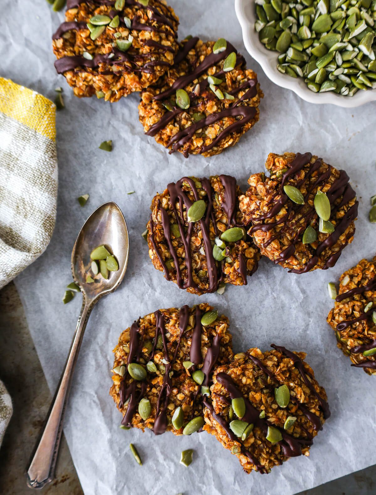 Seven small no bake pumpkin and oatmeal cookies drizzled with chocolate, on parchment, with a spoonfull of pepitas.
