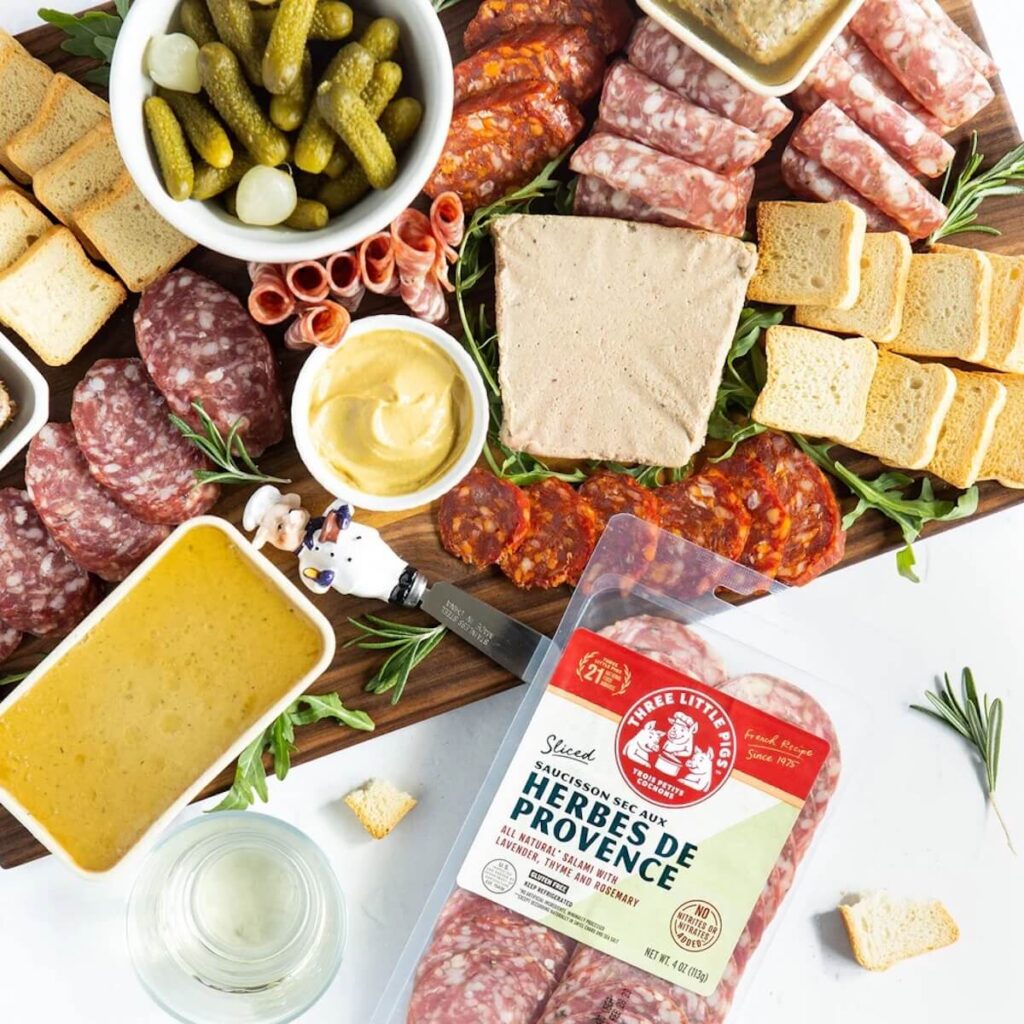 An image of various meeats, cheeses, dips, and garnishes on a charcuterie board with Three Little Picks packaging on the side.