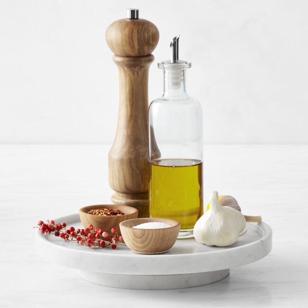 Williams Sonoma's 10 in diameter marble lazy susan placed over a white background with kitchen essentials, such as olive oil, salt and pepper, placed on top of it.