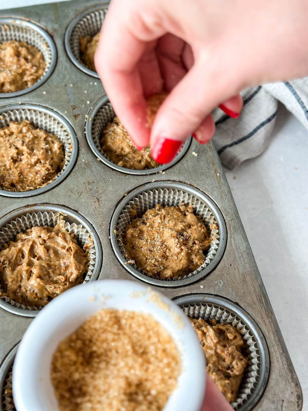 Sprinkling turbinado sugar on top of bran muffin batter in a muffin tin before baking.
