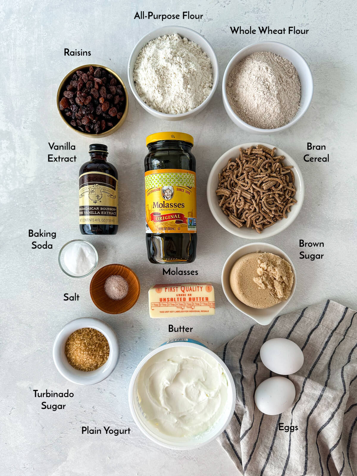 Ingredients for bran muffins in bowls including: flour, whole wheat flour, raisins, bran cereal, molasses, vanilla extract, baking soda, salt, butter, brown sugar, turbinado sugar, plain yogurt, and eggs.