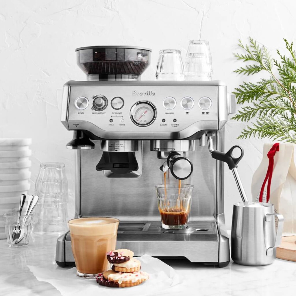 Stainless steel breville barista express espresso machine set in a white kitchen with evergreen tree limbs poking in, making an espresso while Christmas cookies sit off to the side.