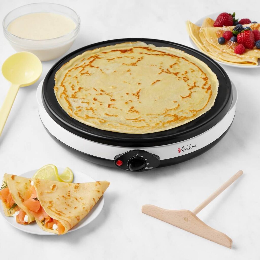 A white euro cuisine crepe maker cooking crepes on a marble surface, with already made sweet and savory crepes and batter close by.