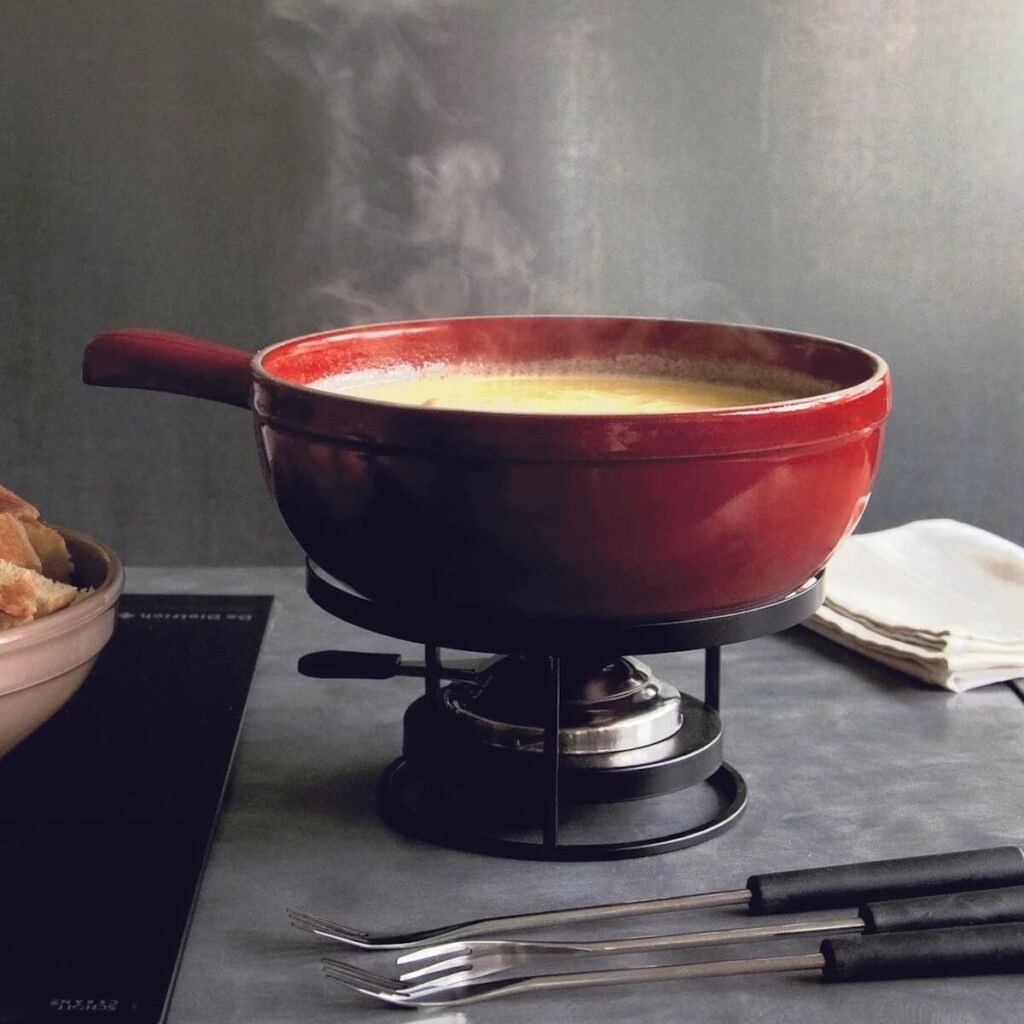 Emily Henry's red fondue pot steaming over a burner, filled with fondue cheese and set in a grey kitchen.