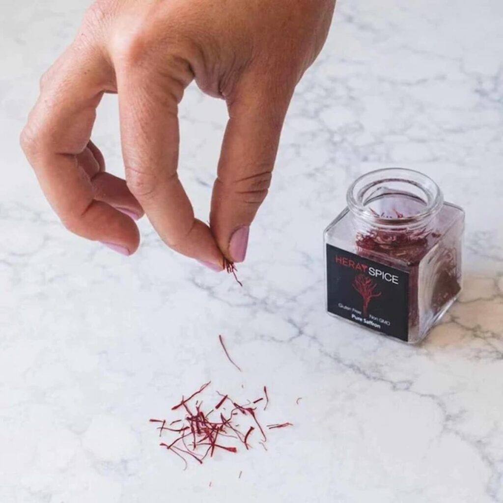 A jar of Heray Spice's red saffron sitting on a marble counter while a hand pinches saffron strands and drops them onto the counter.