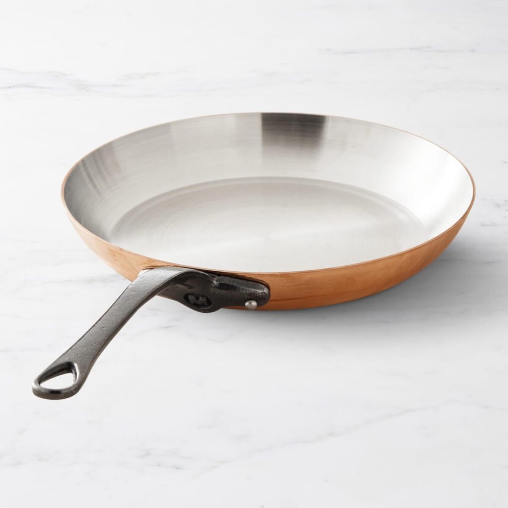 Image of Mauviel's 10 inch polished copper frying pan with a stainless steel interior and bronze handles, sitting on a plain slab of white marble.