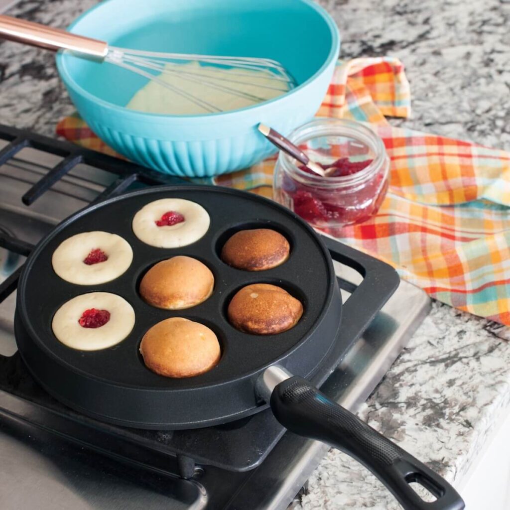 The nordic ware nonstick ebelskiver pan on a gas stove top cooking ebelskivers covered filled with jelly.