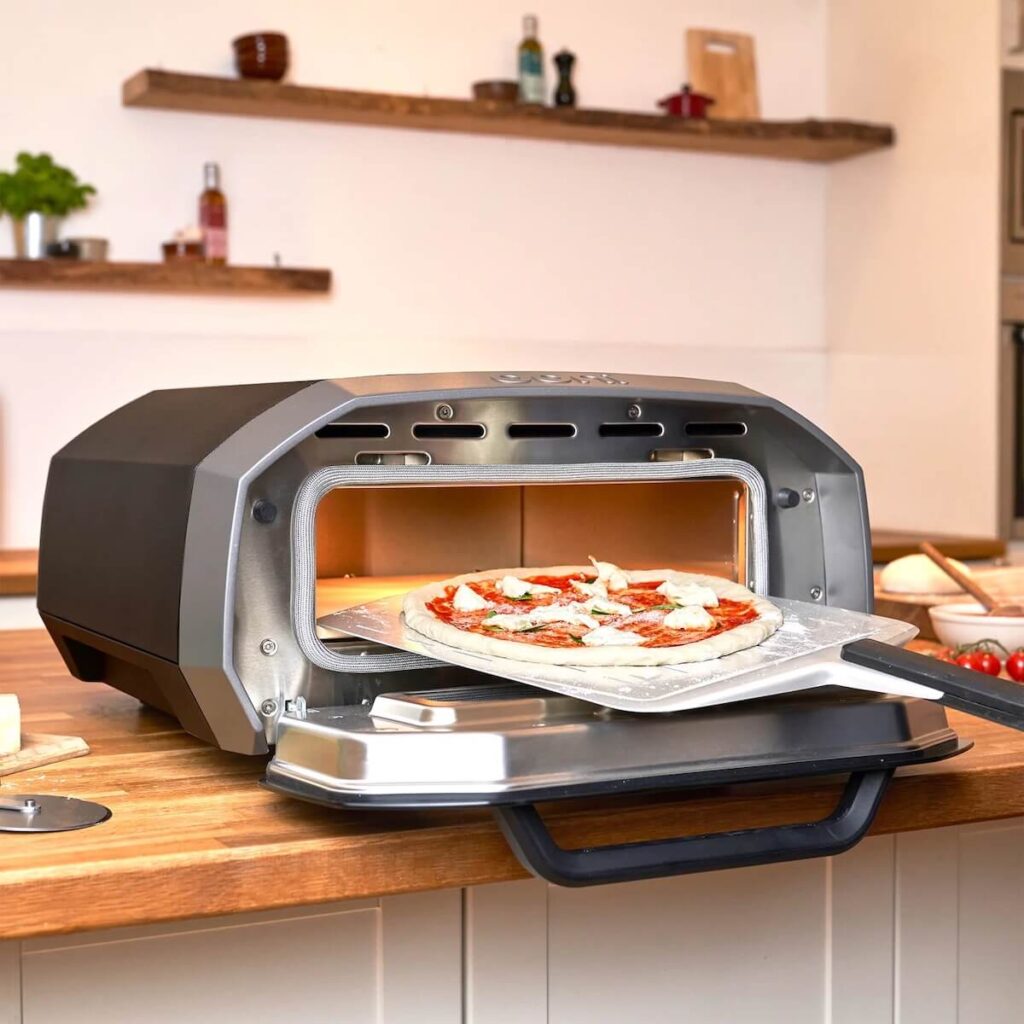 Photo Ooni electric pizza oven in black set inside while a pizza is being placed inside.