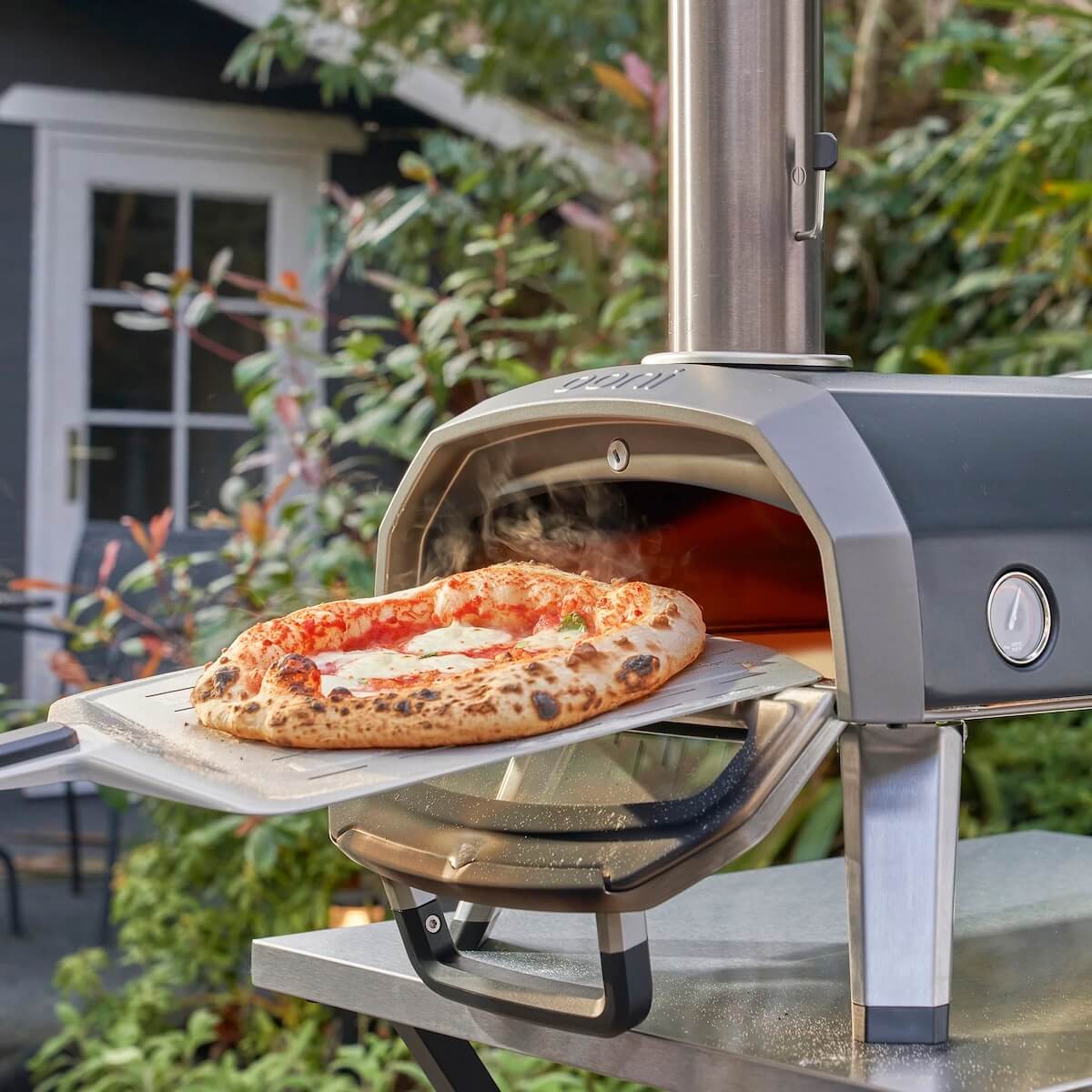 An Ooni Karu Pizza oven being used outside as a user slides a freshly baked margarita pizza out of its fiery inside