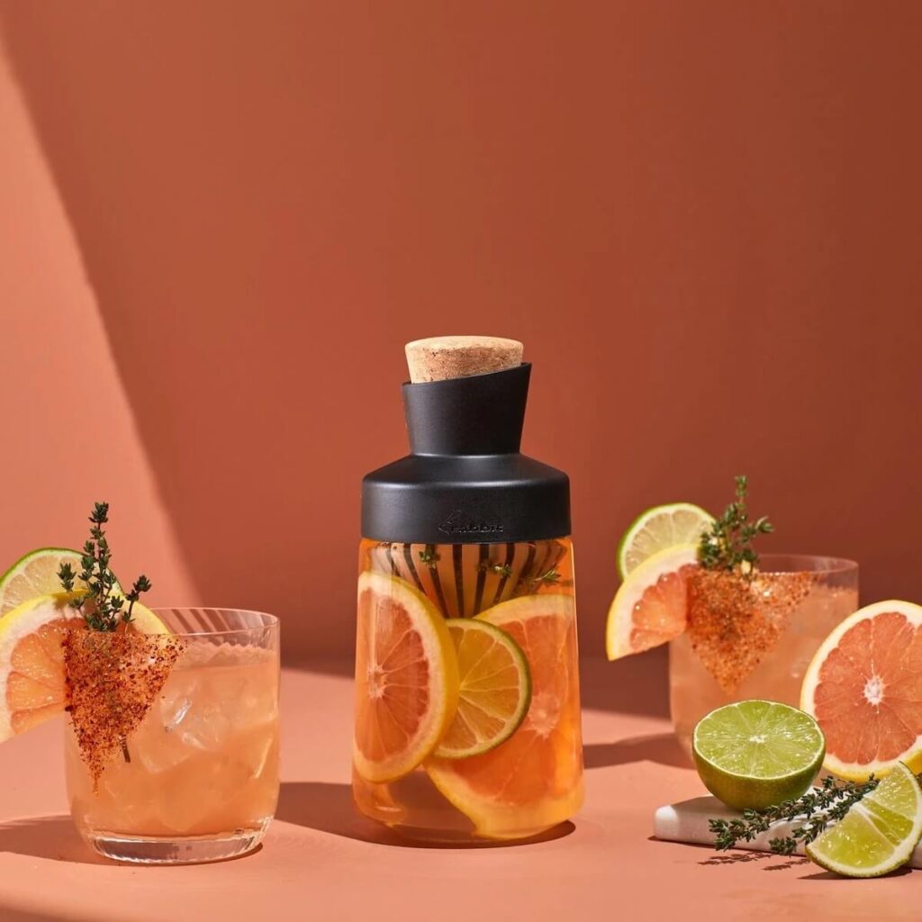 Image of Rabbit's liquor infusing decanter filled with a light pink liquid, slices of grapefruit and lime, and herbs difusing, including a glass with the cocktail inside.
