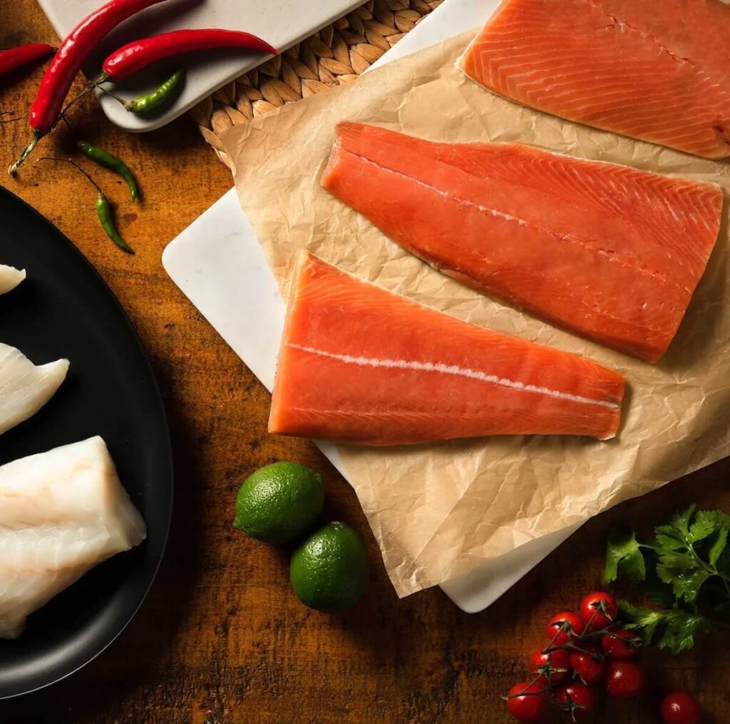 An image of fresh, raw filleys of salmon spread on a wood kitchen counter, next to raw white fish fillets and products.