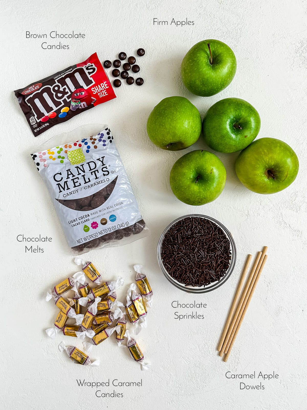 Ingredients for spiderweb caramel apples, including chocolate candies, chocolate melts, caramels, apples, sprinkles and caramel apple dowels.