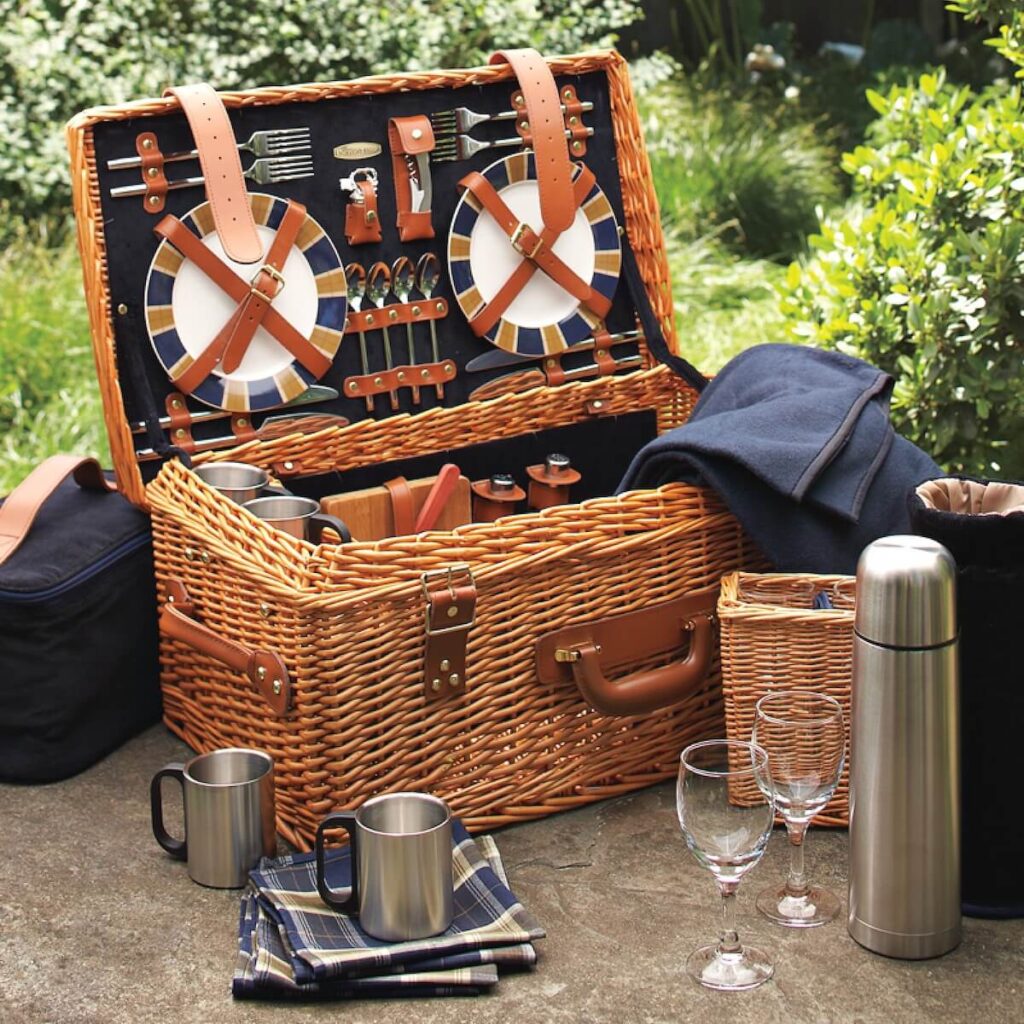 Outdoor scene of William Sonoma's wicker picnic basked, opened, displaying a place set inside. including procelain plates and hand blown wine glasses.