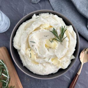 Overhead view of a grey bowl of rosemary mashed potatoes with fresh rosemary garnish and melted butter.