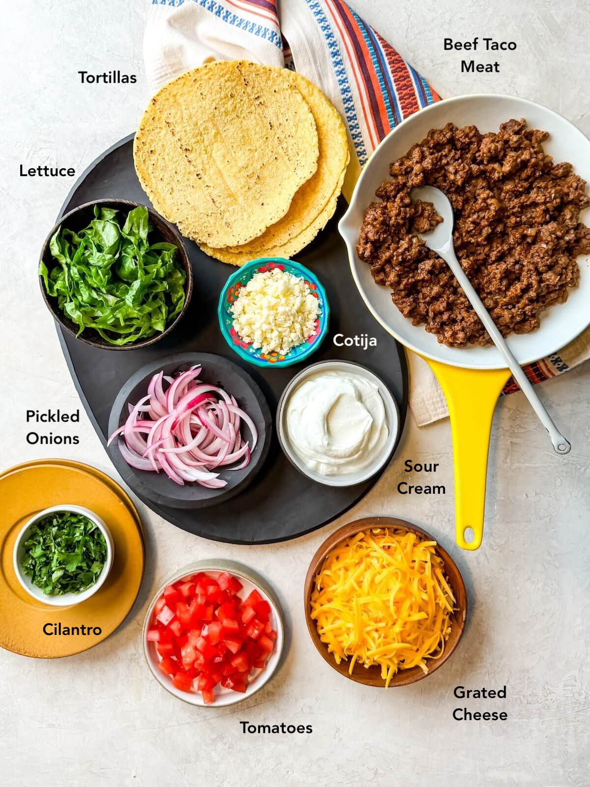 Ground beef tacos ingredients on a platter including taco meat, tortillas, lettuce, red onion, cotija, sour cream, cheese, tomatoes, and cilantro.