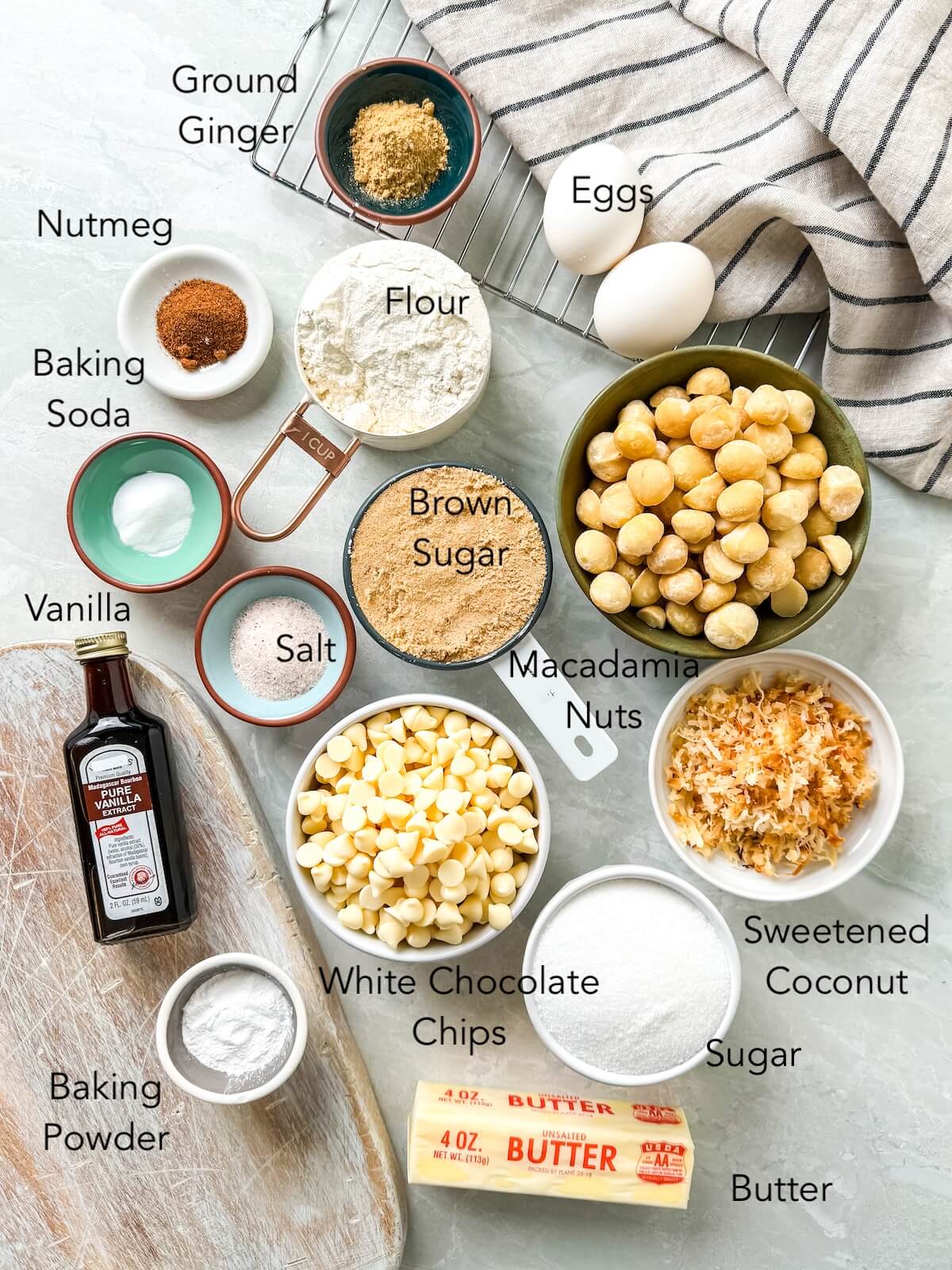 Ingredients for macadamia nut cookies on a kitchen surface including: flour, ground ginger, nutmeg, baking soda, baking powder, salt, sugar, brown sugar, vanilla, eggs, coconut, butter, white chocolate chips and macadamia nuts.