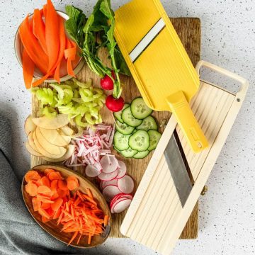 Sliced vegetables and fruit on a cutting board with two different mandoline slicers.