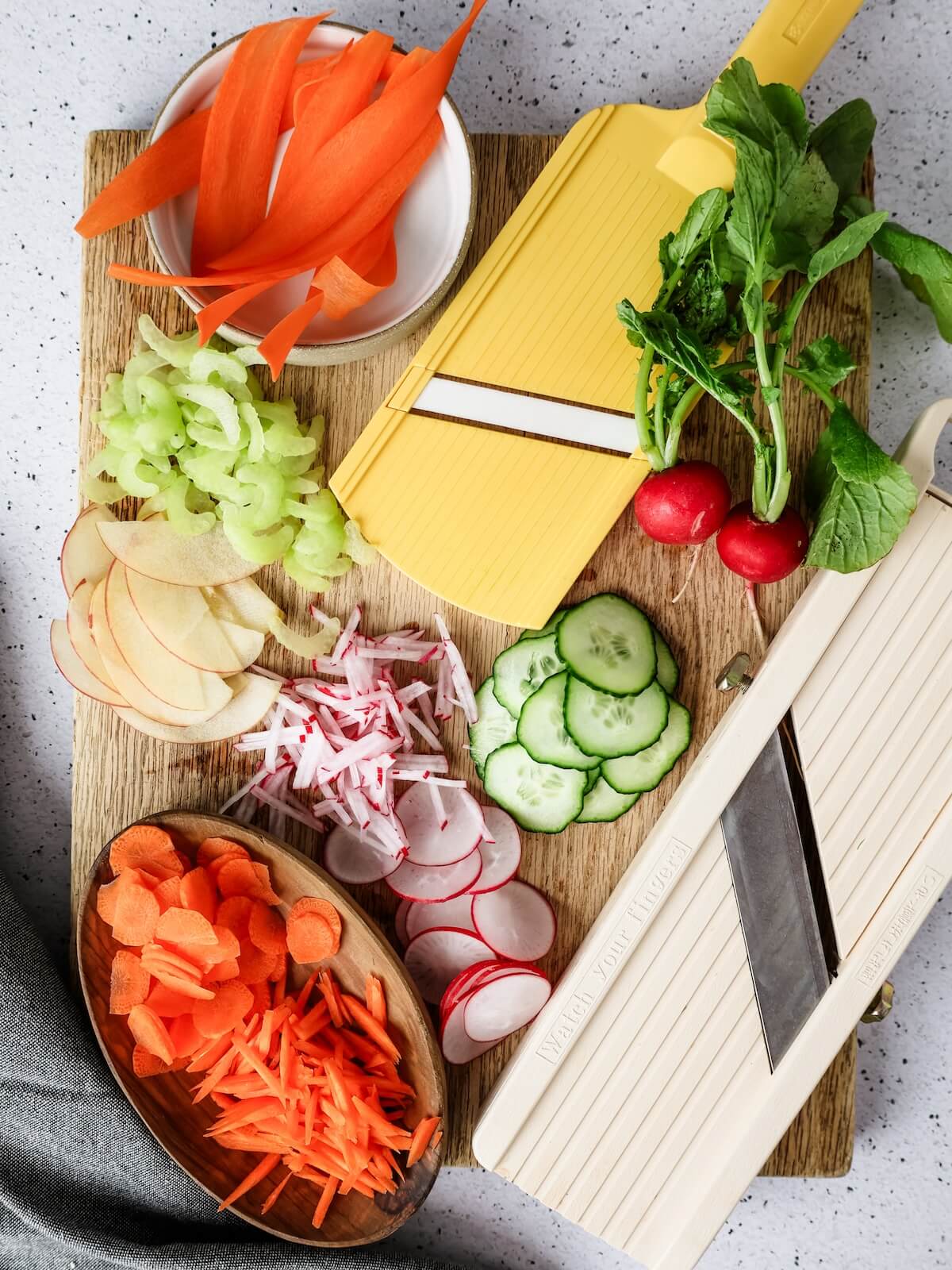 Sliced vegetables and fruit on a cutting board with mandoline slicers.