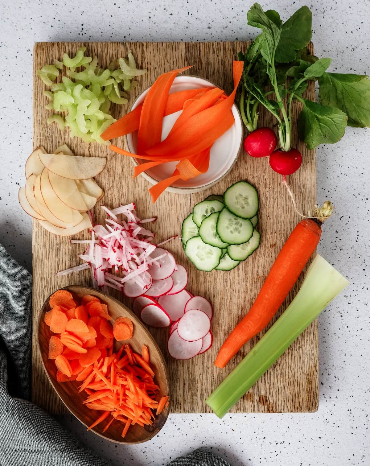 Sliced vegetables and fruit on a cutting board that have been cut with a mandoline slicer.