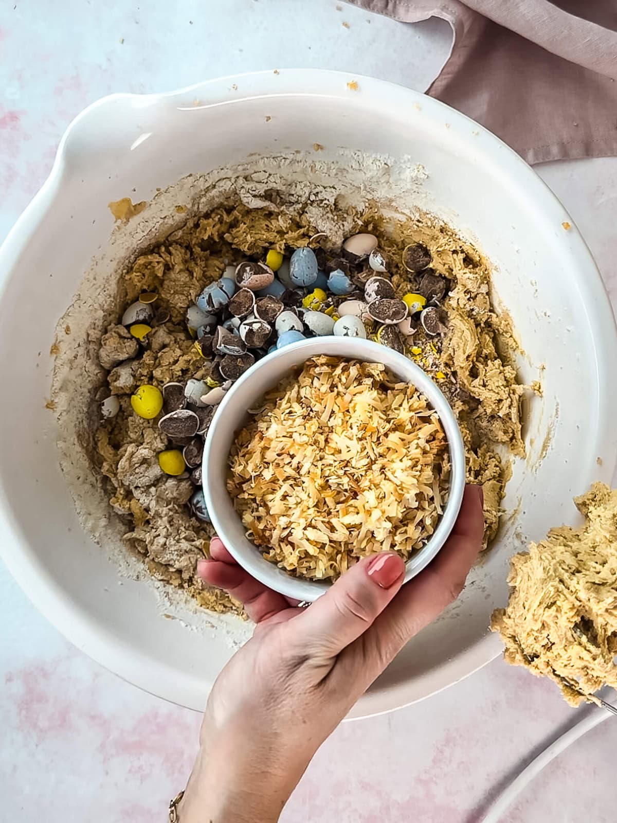 Hand adding toasted coconut to a bowl of blondie batter in a white mixing bowl.