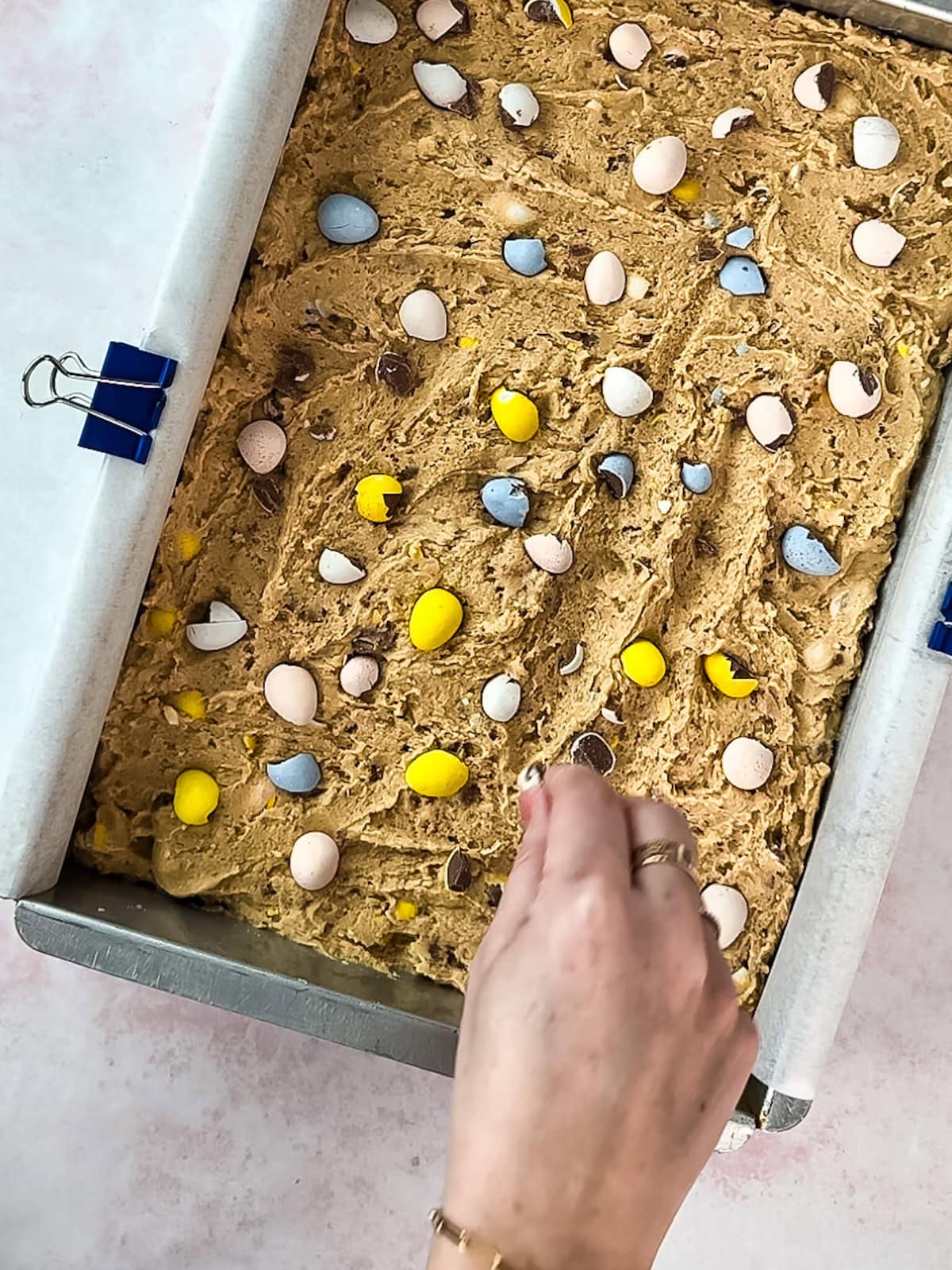 Hand sprinkling crushed mini eggs on top of blondie dough in a baking pan.