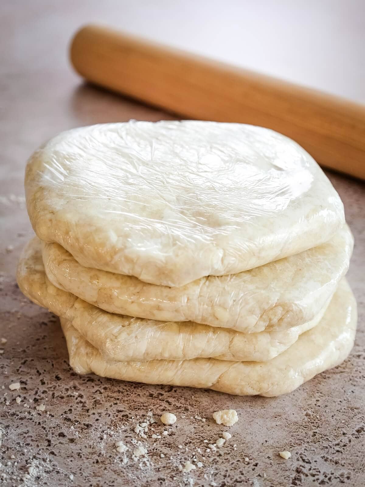 Four plastic wrapped discs of pie crust with a rolling pin in the background.