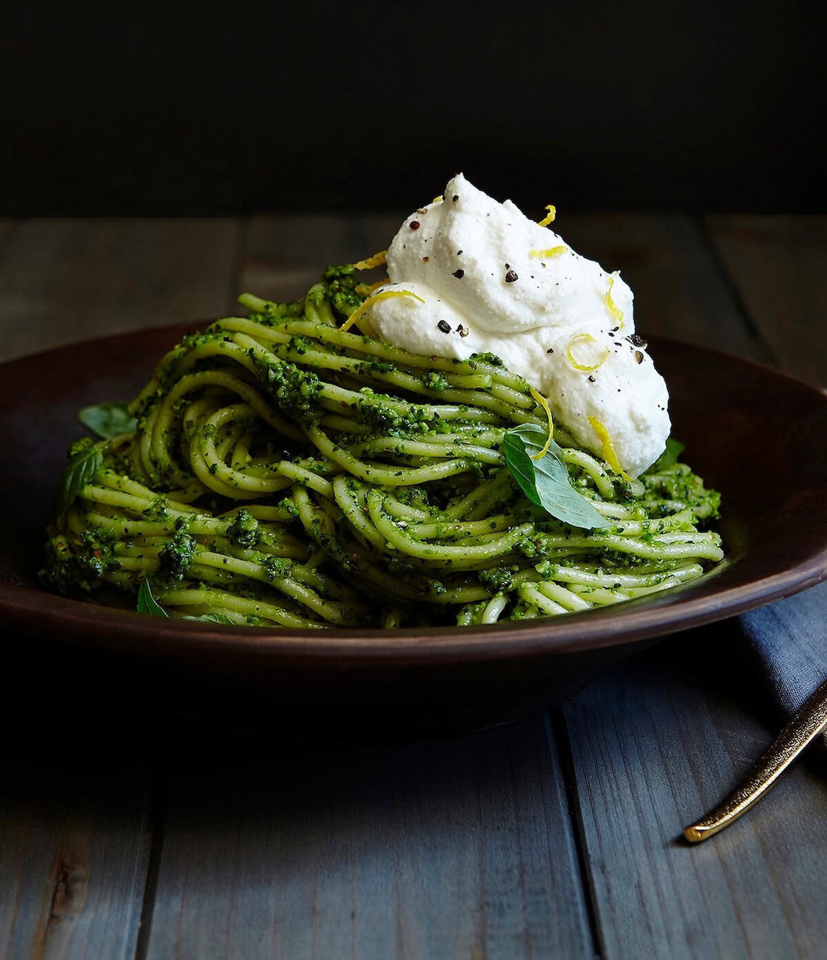 pesto pasta with lemon ricotta in dark bowl with black background and basil leaves.