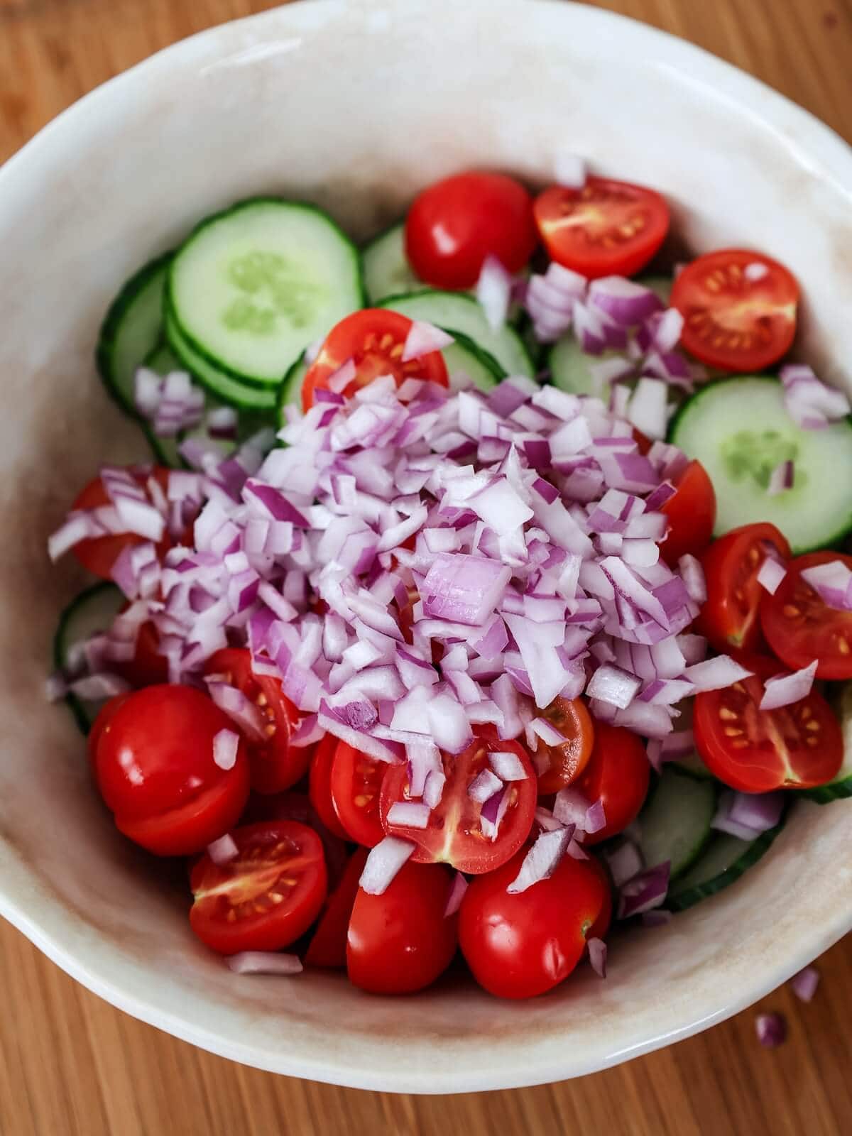 making cucumber salad in a bowl with red onion, tomatoes and cucumber.