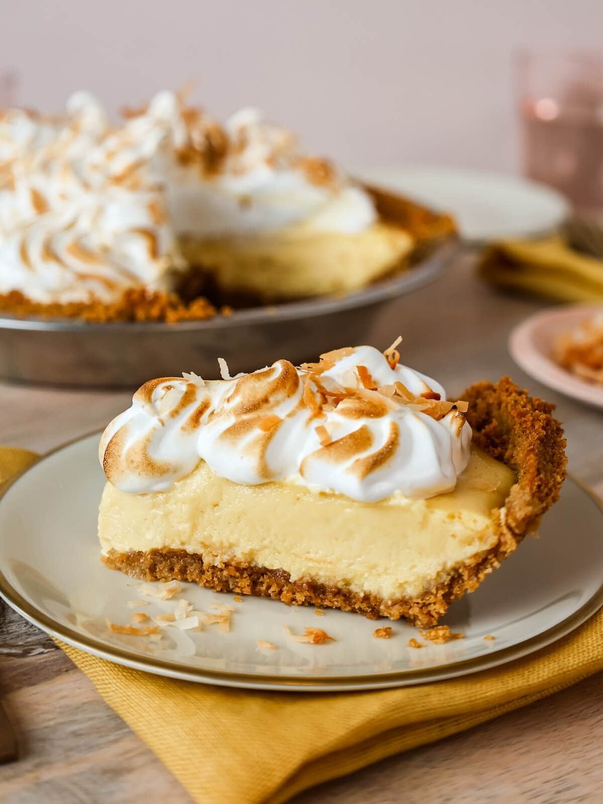Close-up of a slice of mango meringue pie with toasted coconut on a plate, with the full pie in the background.