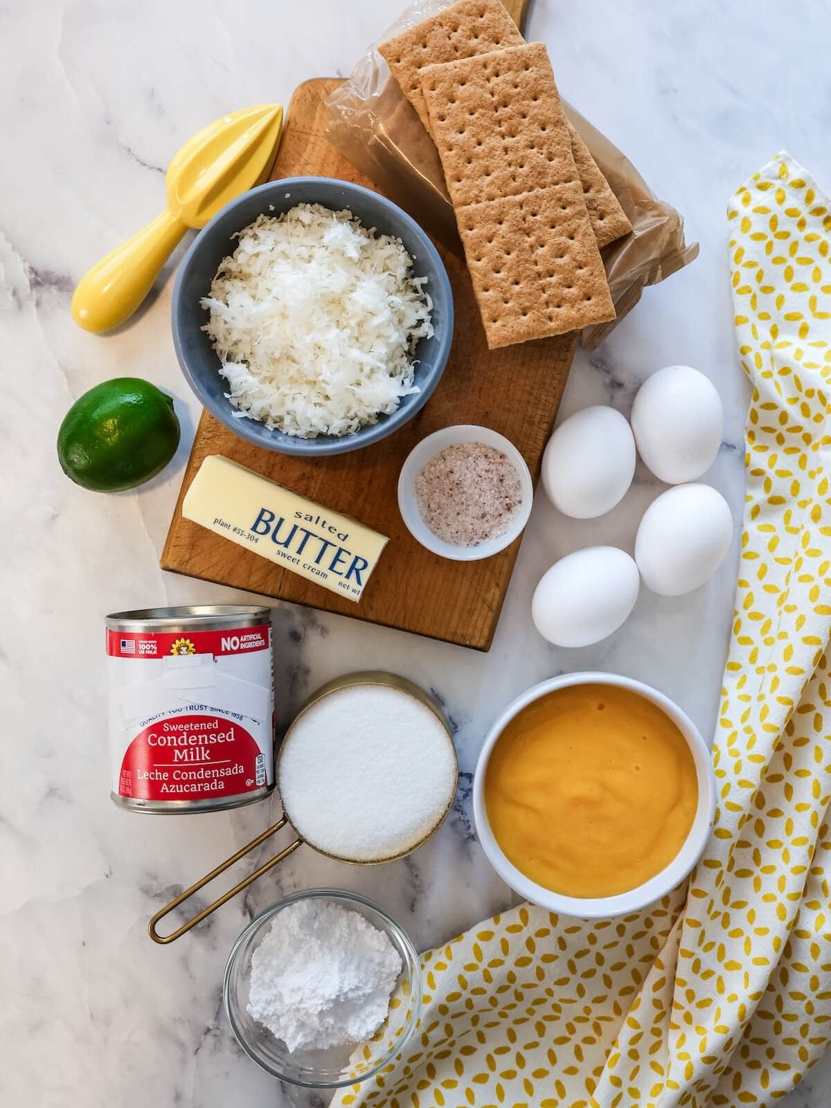 Flat lay of ingredients for mango pie, including eggs, graham crackers, shredded coconut, lime, butter, sugar, condensed milk, and mango puree.