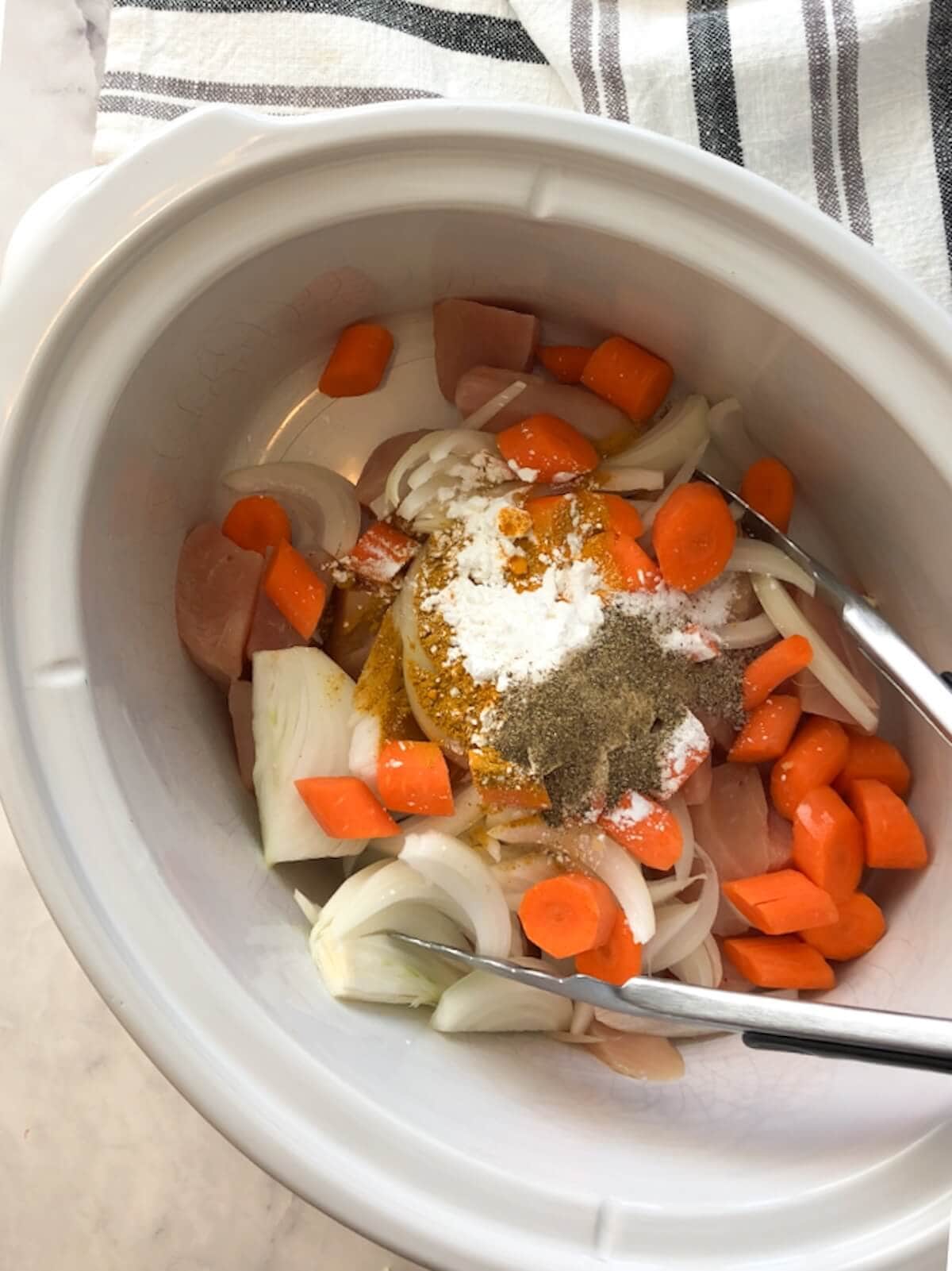 Ingredients including chicken, carrots, and onions placed in a slow cooker with seasonings sprinkled on top.