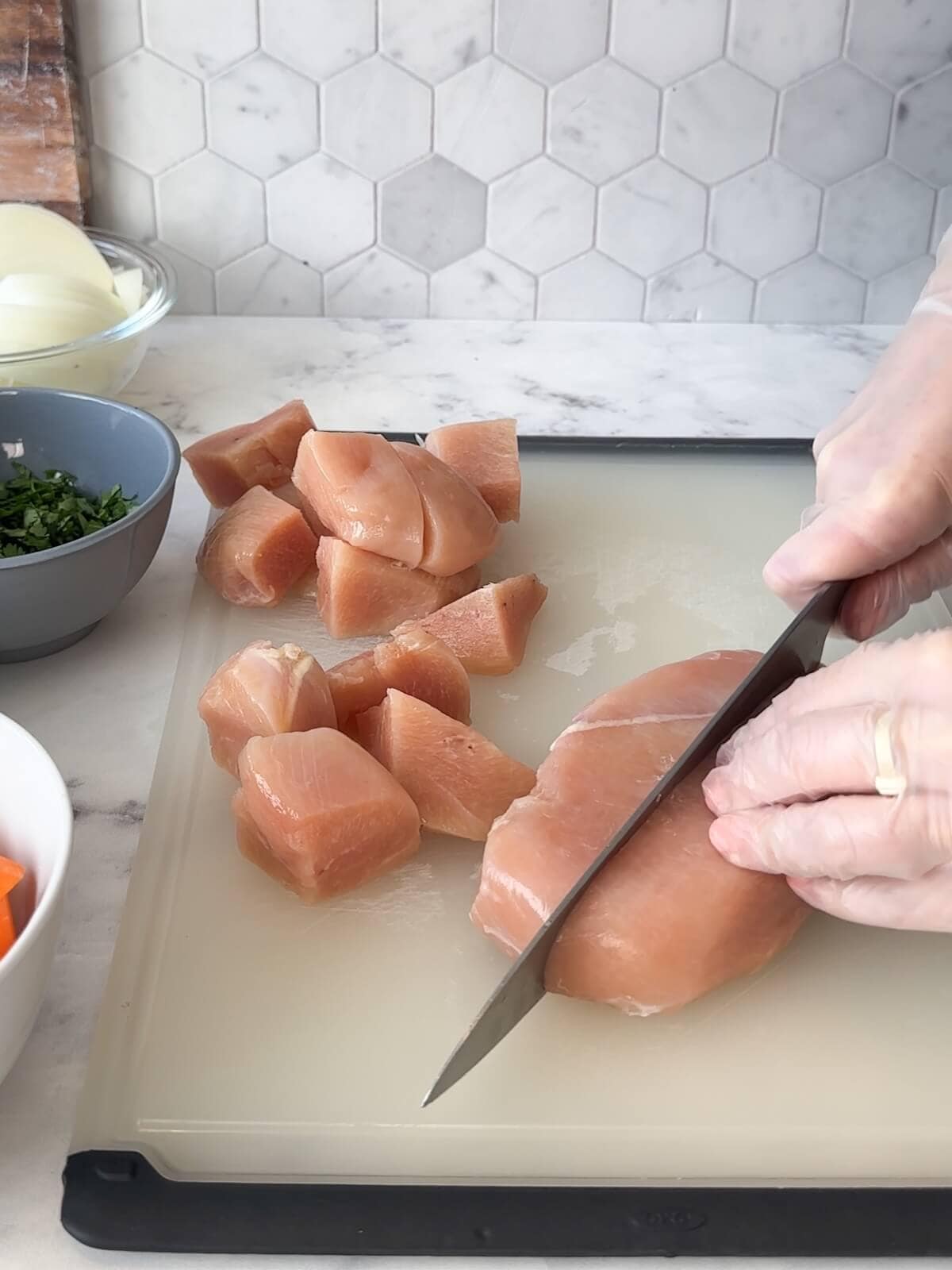 Chopping raw chicken breast into chunks on a cutting board, surrounded by bowls of prepped vegetables.