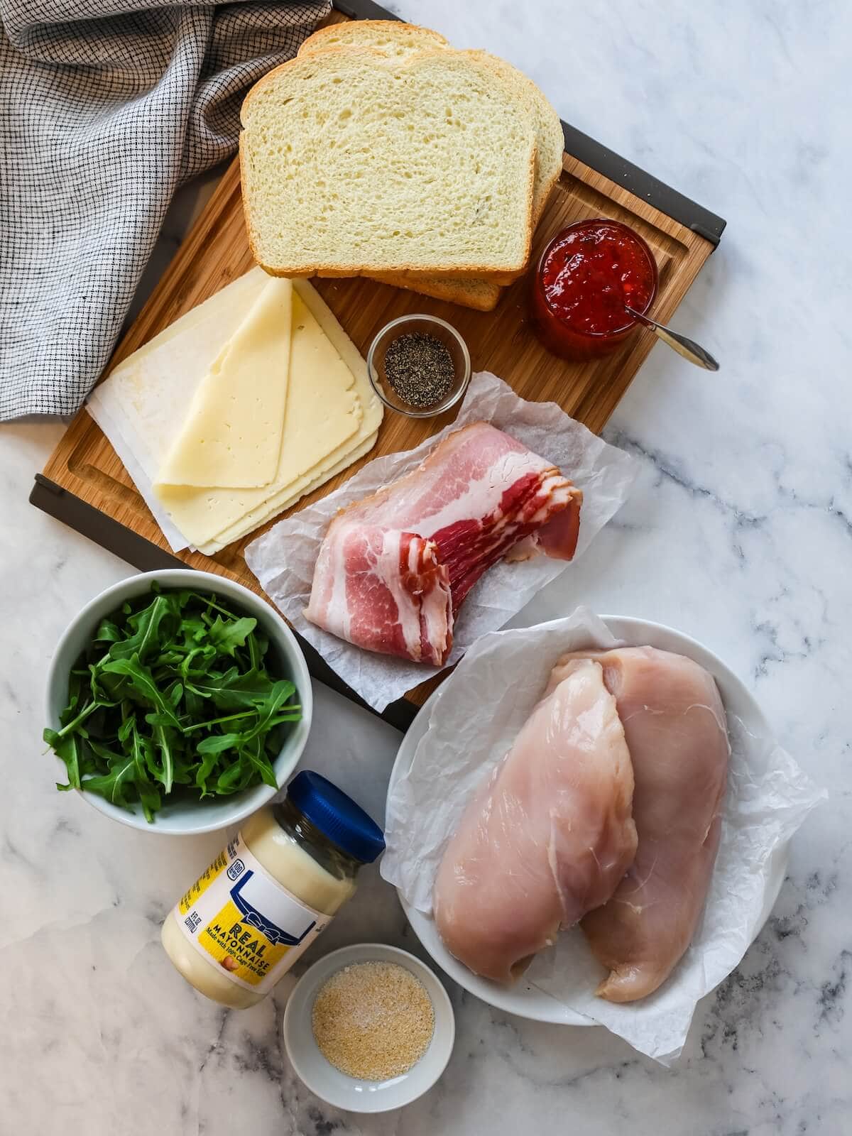 ingredients for sweet and spicy chicken sandwich including bread, cheese, sweet pepper jelly, salt and pepper and garlic powder, chicken, mayo, arugula, and bacon.