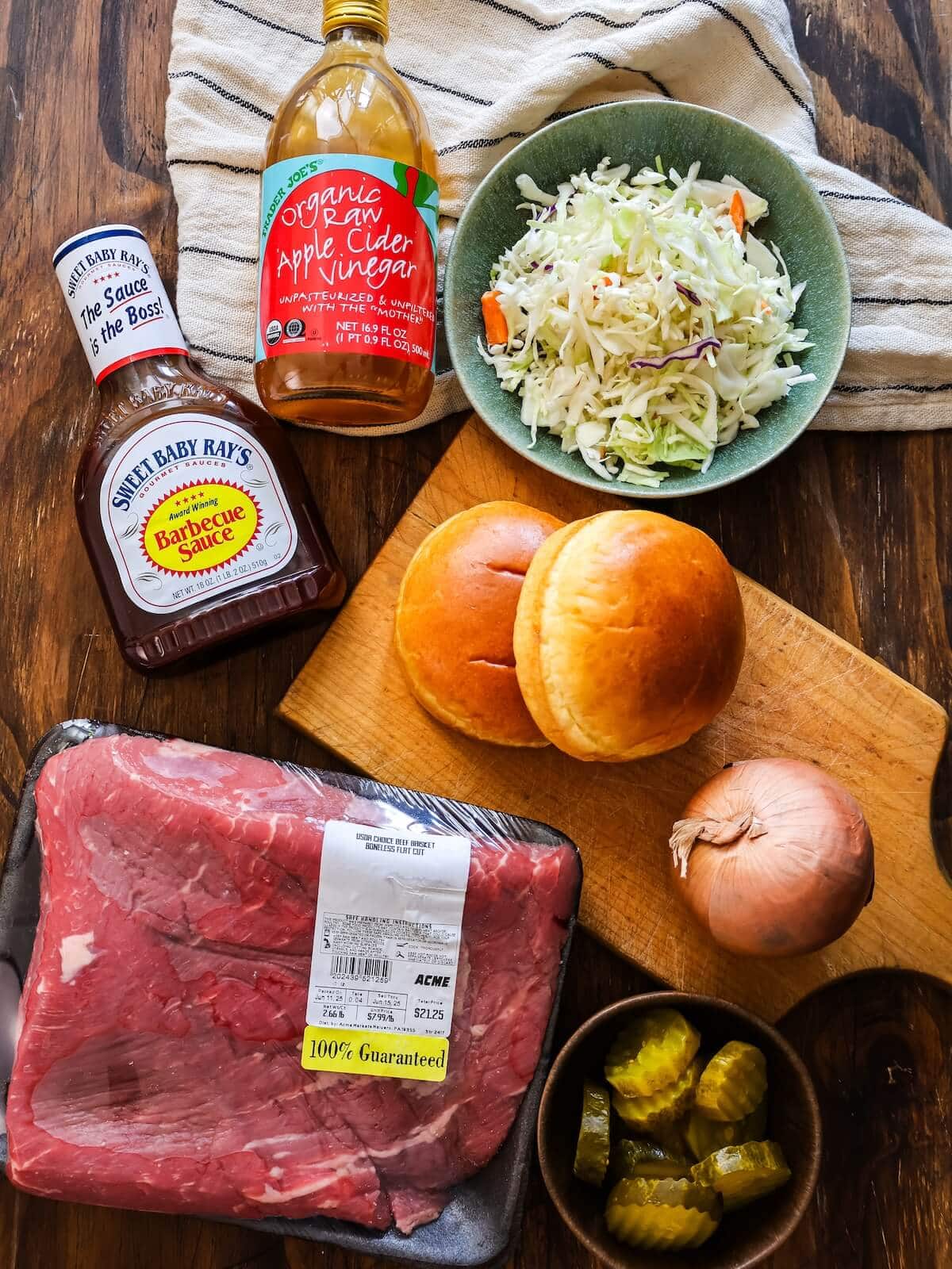 ingredients for slow cooker brisket sandwiches including BBQ sauce, apple cider vinegar, coleslaw, onion, soft rolls, and pickles.