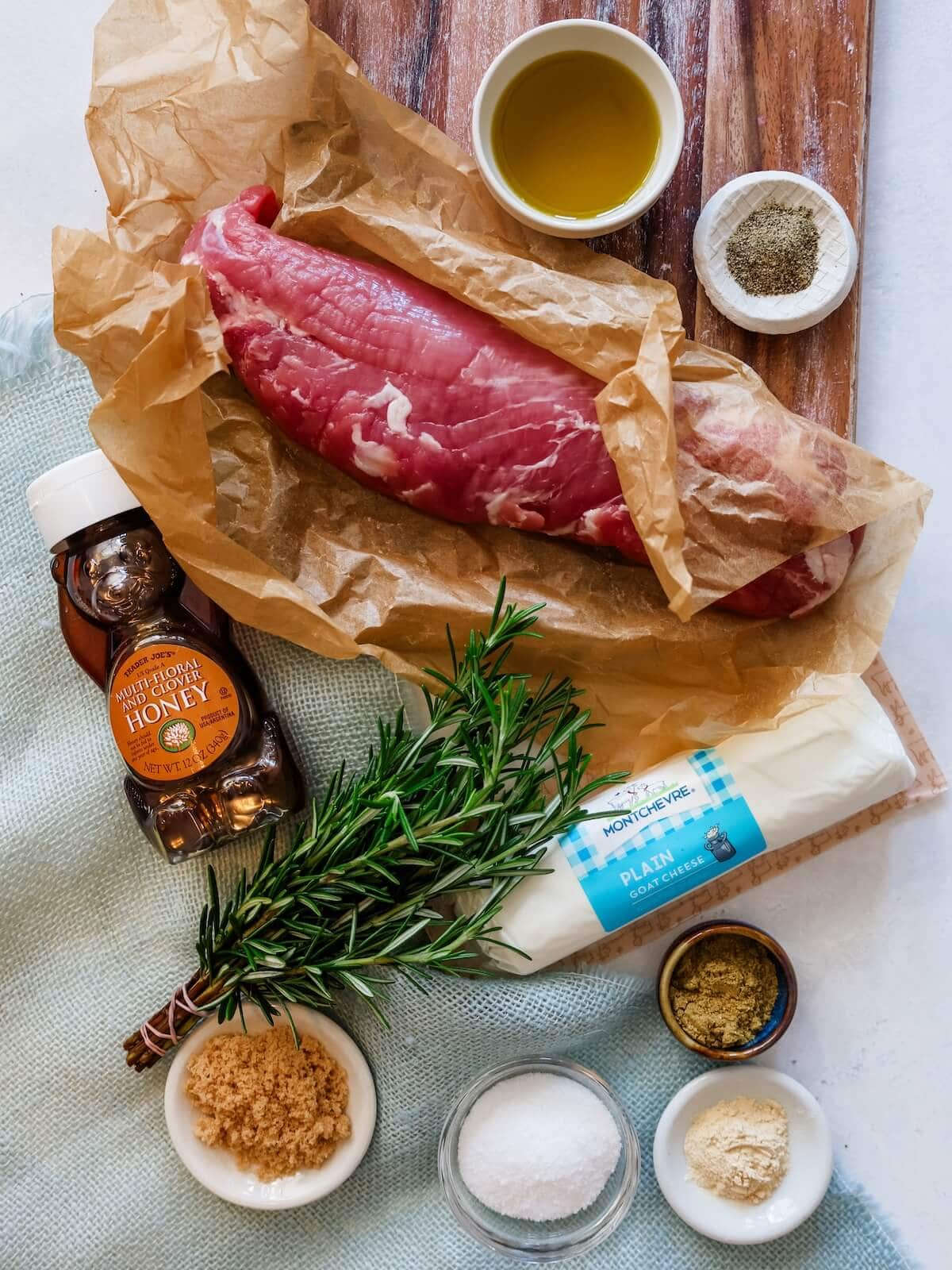 Ingredients for grilled pork tenderloin on a cutting board including pork, oil, pepper, salt, garlic powder, coriander, brown sugar, honey, goat cheese and rosemary.