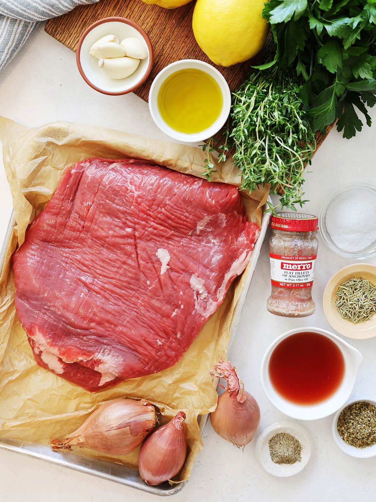 Ingredients for grilled flank steak with salsa verde including steak, garlic, oil, lemons, herbs, dried herbs, shallots, vinegar, salt, and anchovies.