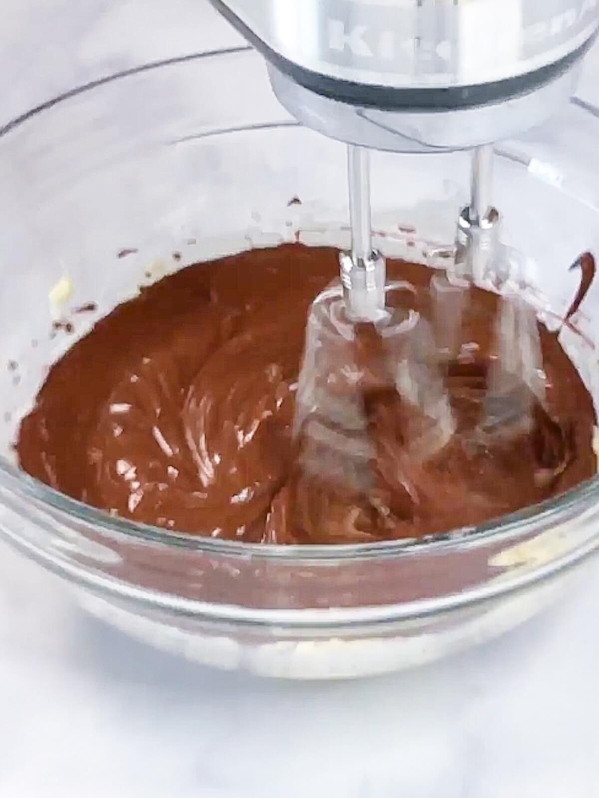 Using a hand mixer to blend in cholate mixture to butter mixture.