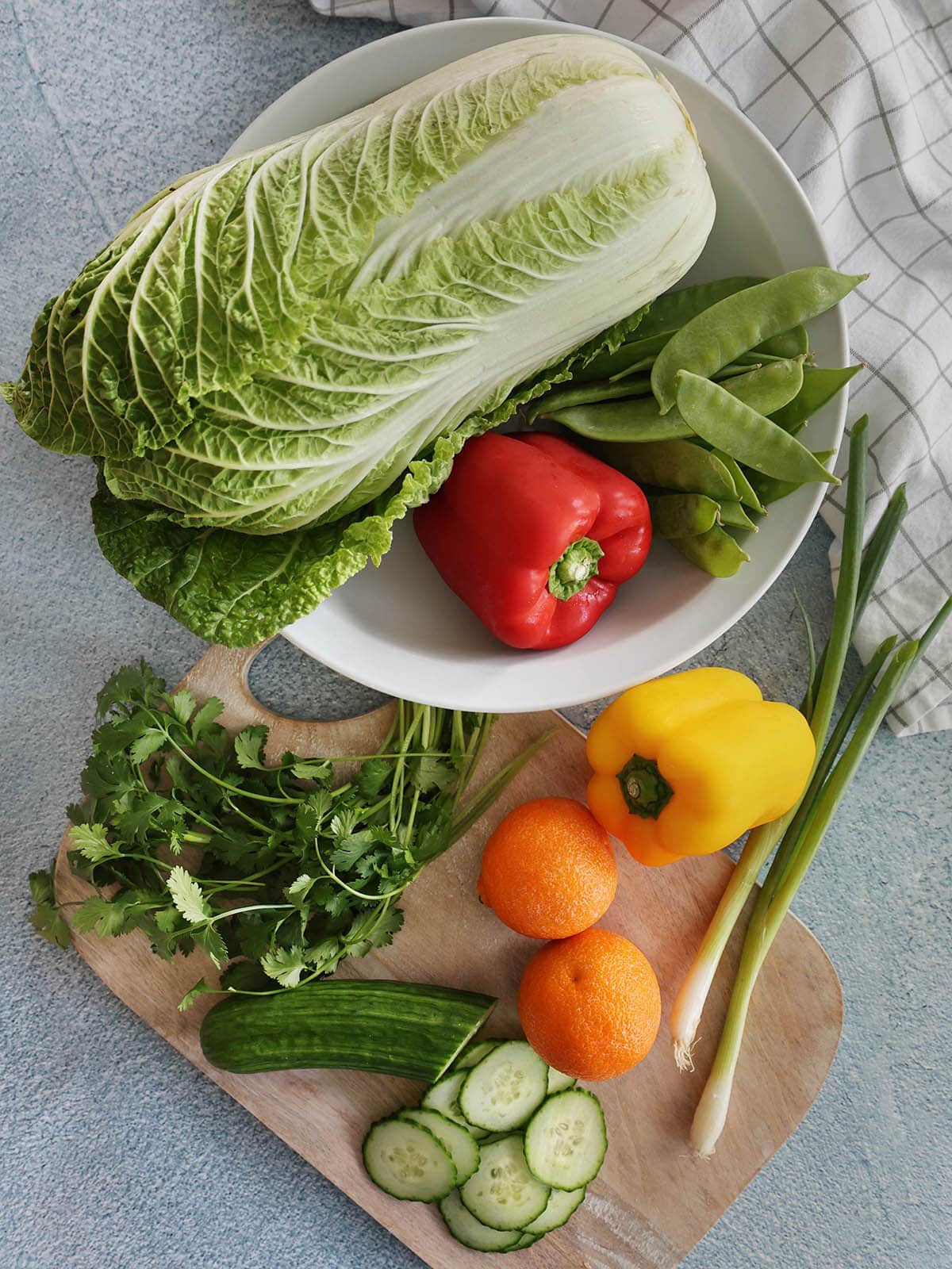 Ingredients for an asian slaw including napa cabbage, bell peppers, snow peas, cilantro, cucumber, scallion, and clementines.