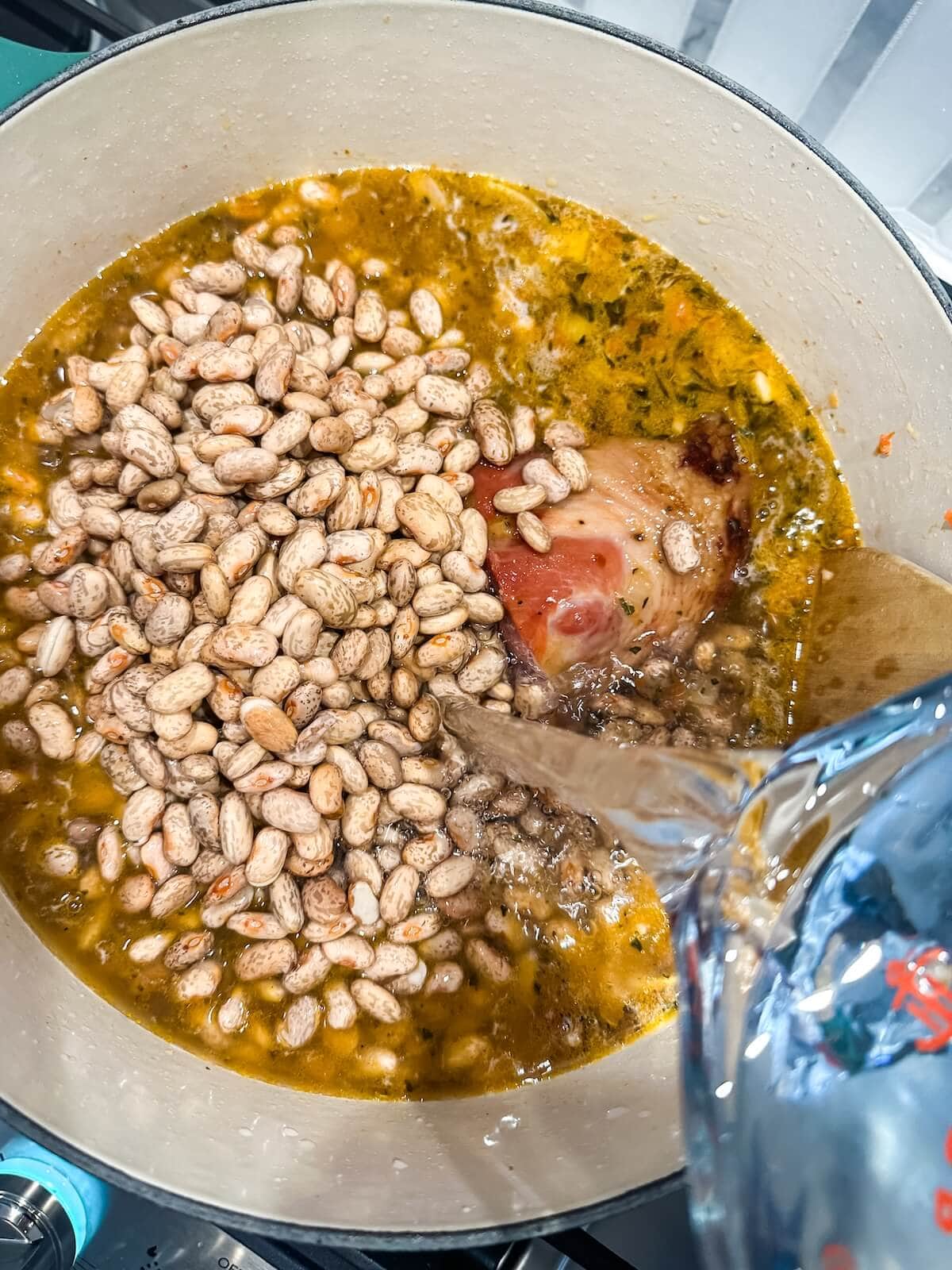 Adding soaked beans, ham hock, water, and broth to pot for frijoles charro.