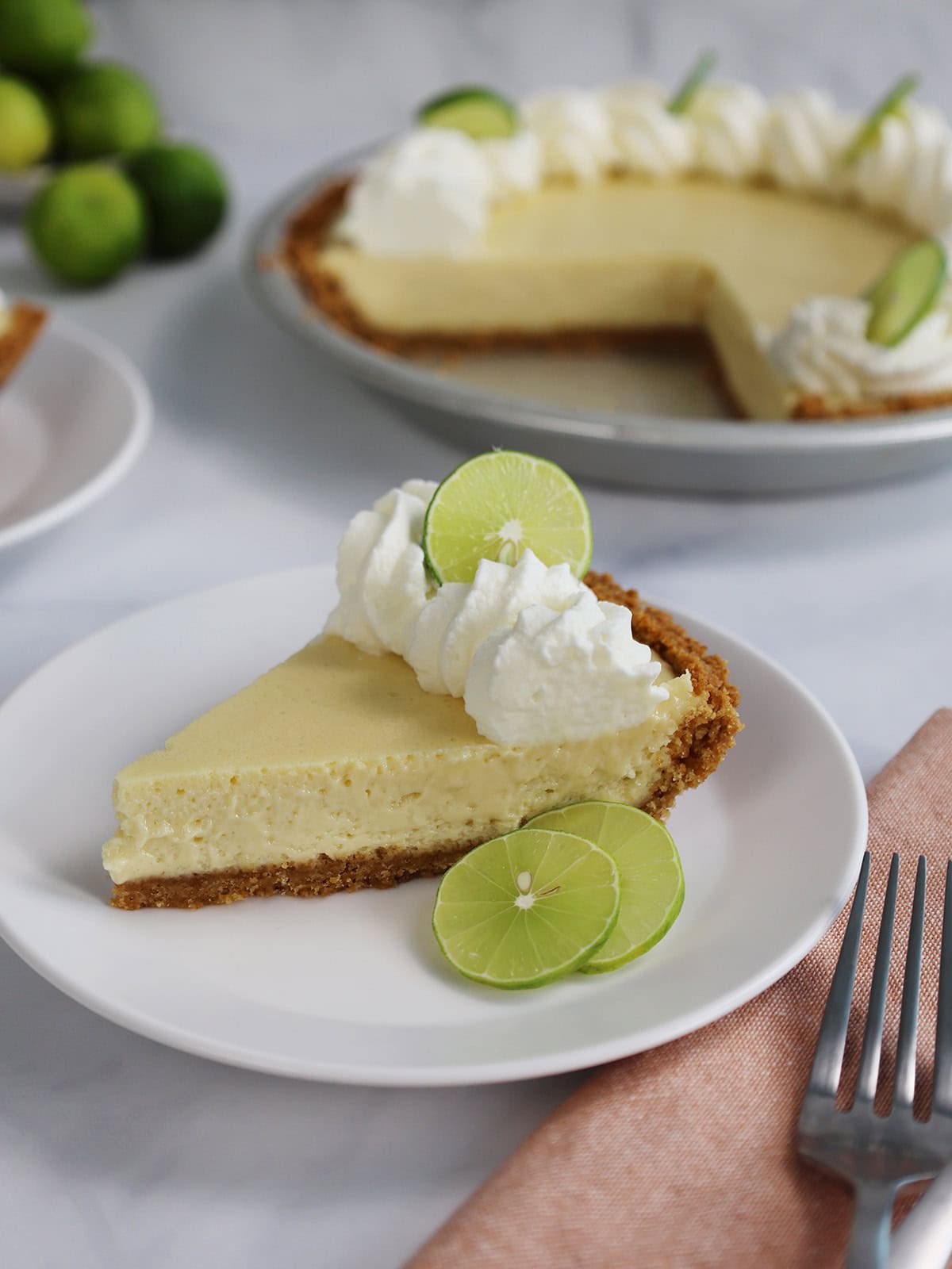 Side view of a slice of Key lime pie on a white plate with the rest of the pie in the background.