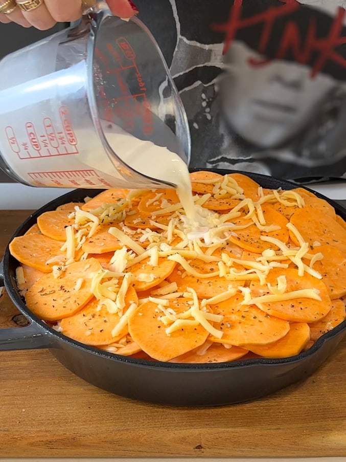 Pouring cream mixture over sweet potatoes in a cast iron baking dish.