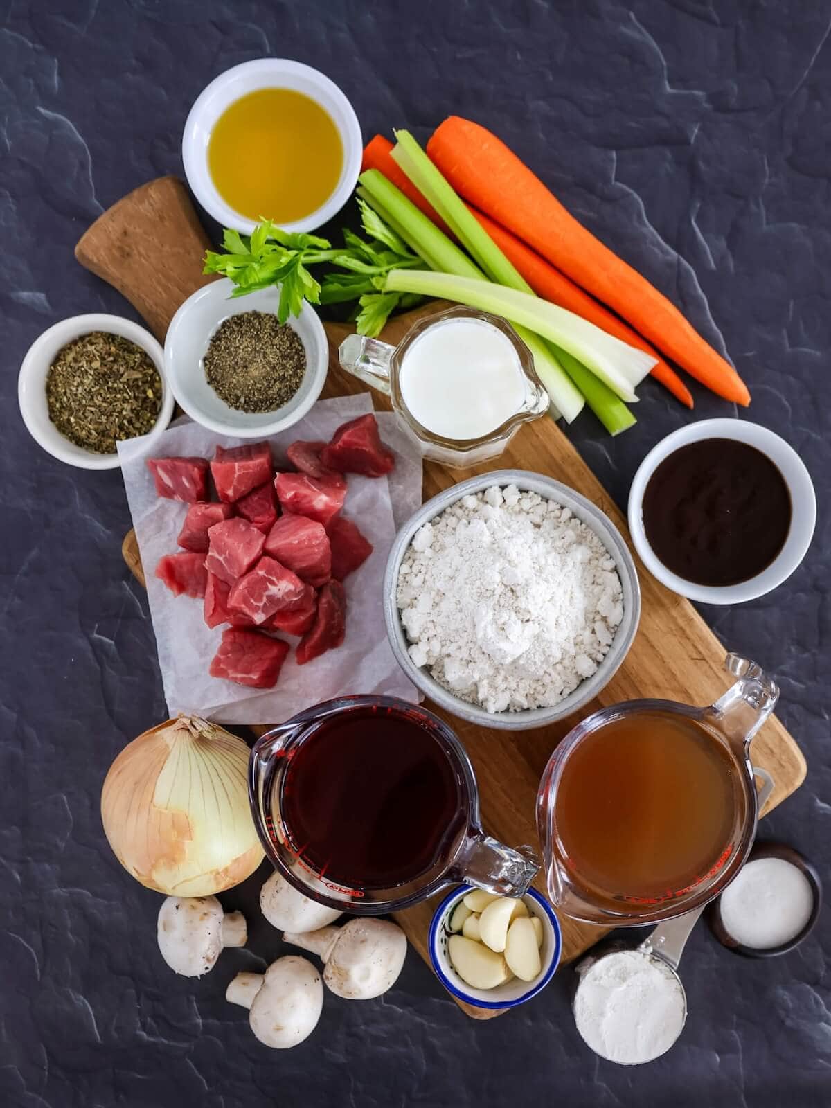 Ingredients for beef stew with biscuit topping including flour, salt and pepper, stew meat, oil, carrots, celery, onion, garlic, mushrooms, wine, beef broth, steak sauce, Italian seasoning, biscuit mix, and milk.
