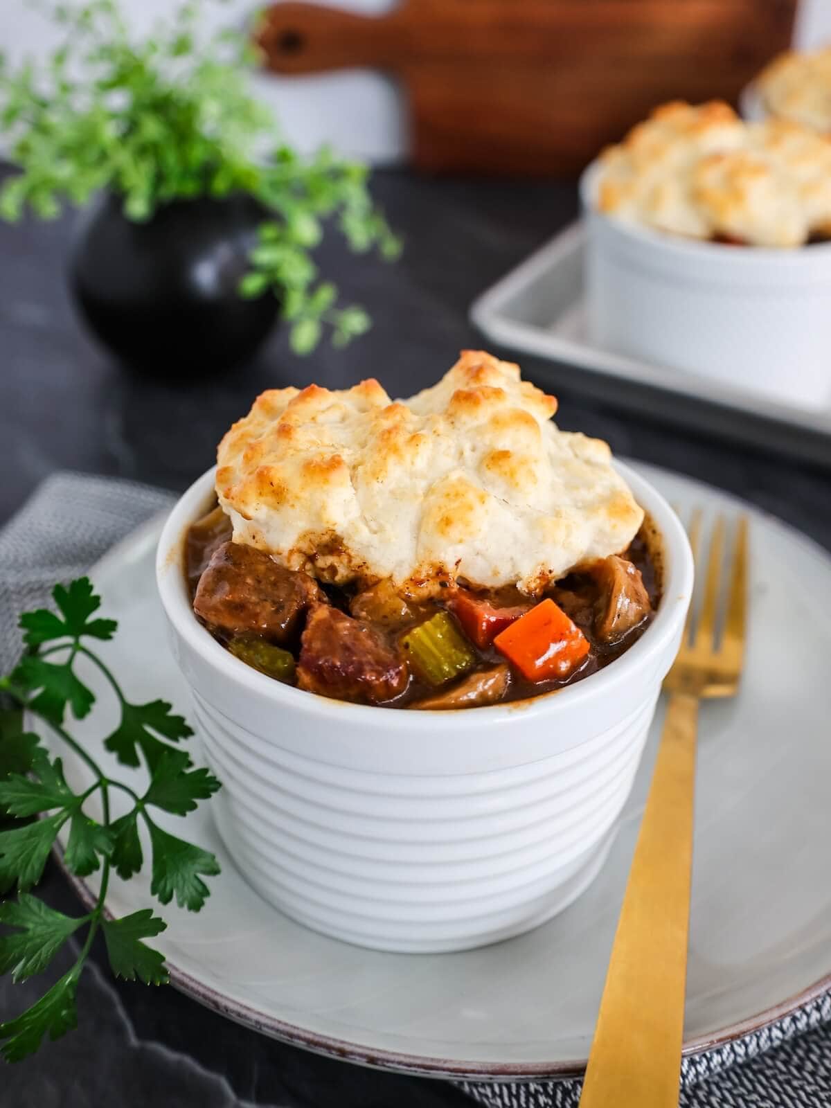 A bowl of beef stew with biscuit topping on a white plate next to a gold fork.