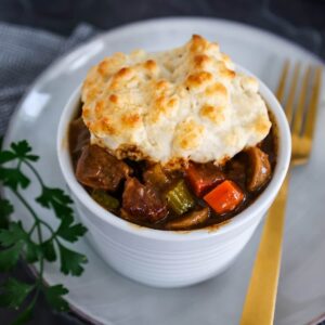 Close up of a bowl of beef stew with biscuit topping on a white plate next to a gold fork.