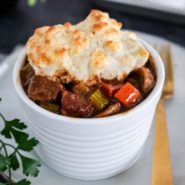 Close up of a bowl of beef stew with biscuit topping on a white plate next to a gold fork.