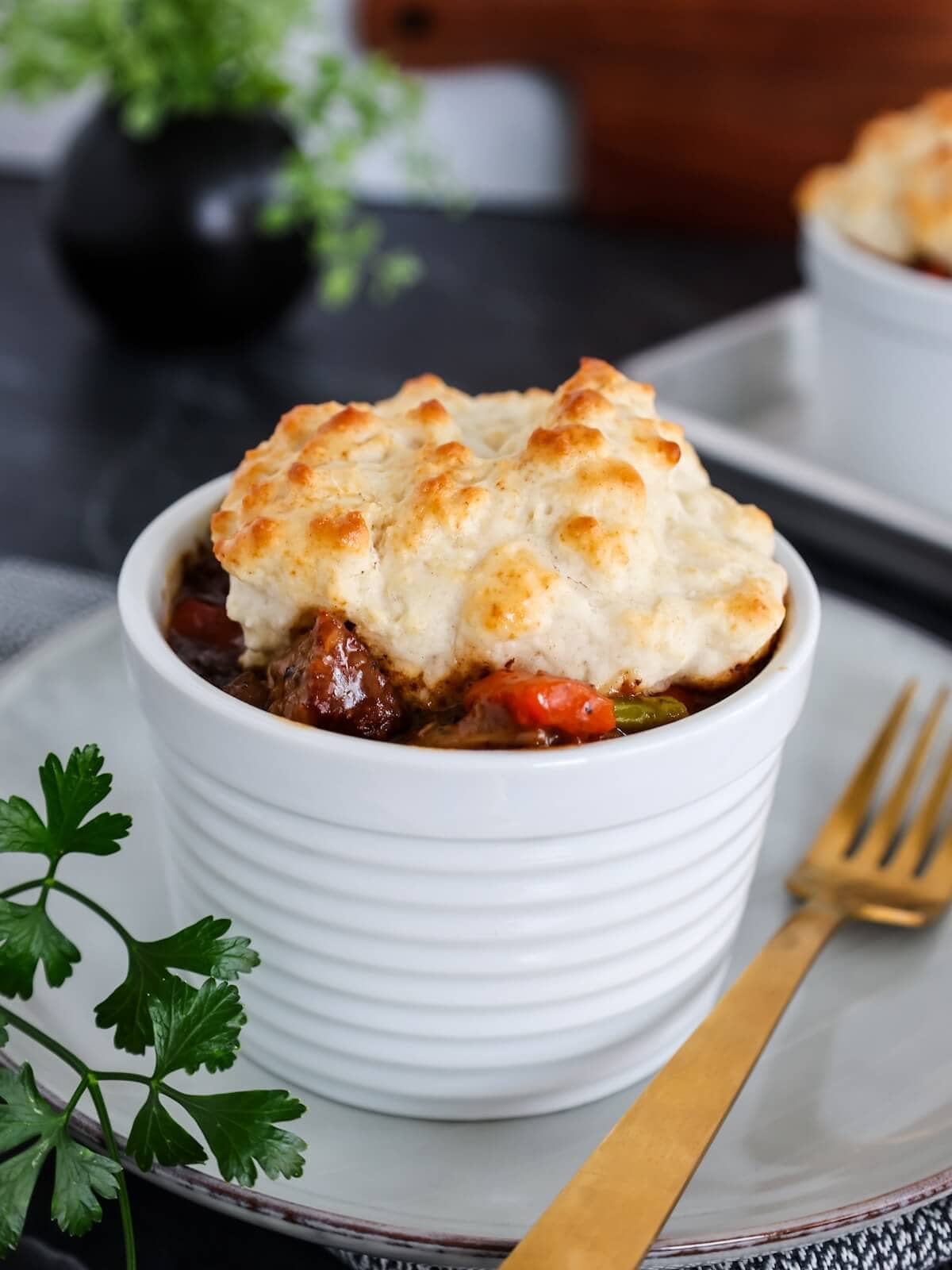 Close up of a bowl of beef stew with biscuit topping on a white plate next to a gold fork.