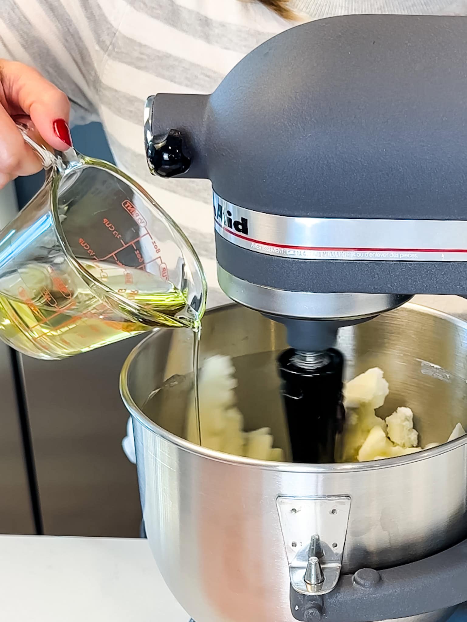 Hand pouring oil into a mixing bowl with creamed butter and sugar in it.