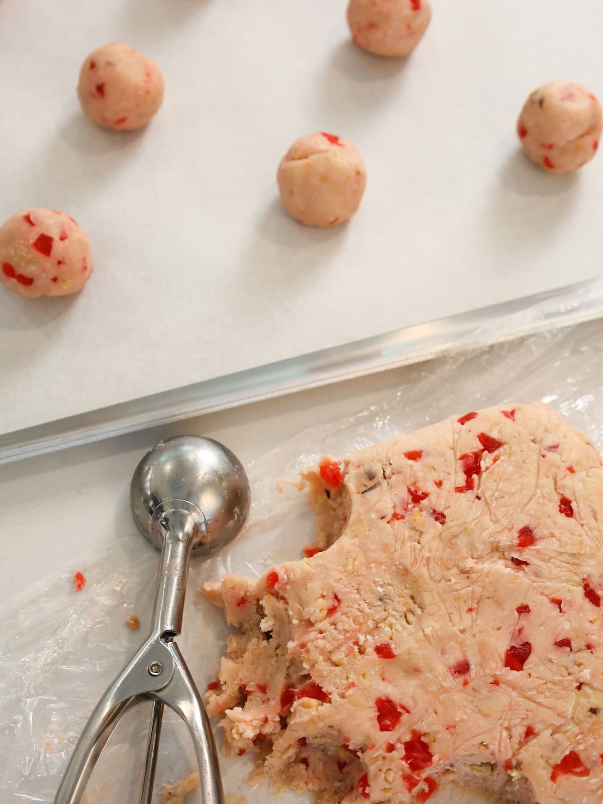 A slab of cookie dough next to a cookie scoop next to a baking sheet with cookie dough balls on it.