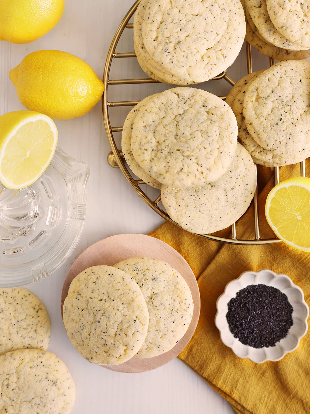 Top view of lemon poppyseed cookies on a golden wire rack and scattered on a small plate and countertop next to lemons.